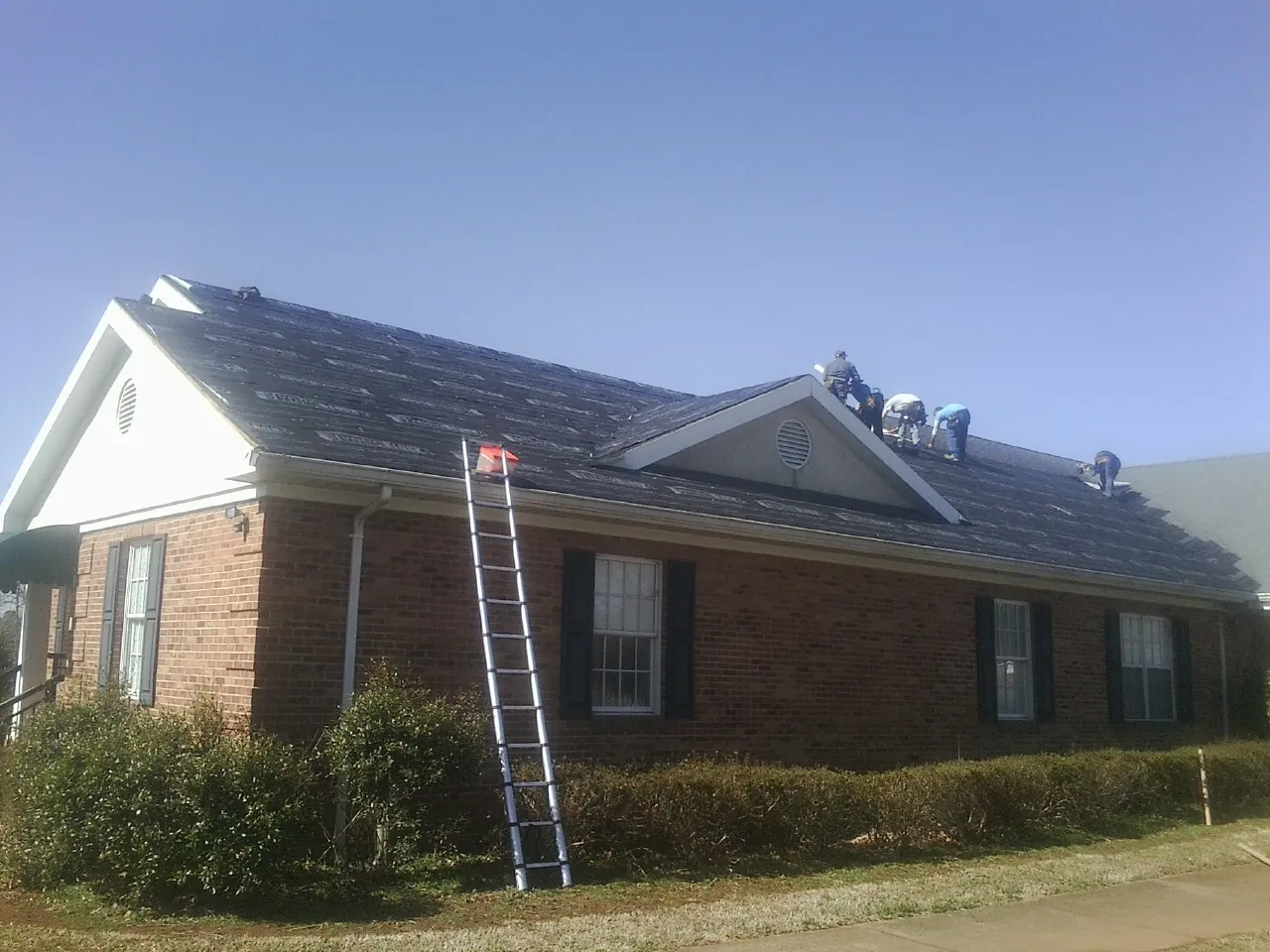 Crew tearing shingles off a brick ranch roof with a ladder set — Emergency Roof Repair work by our crew in Mc Clellanville