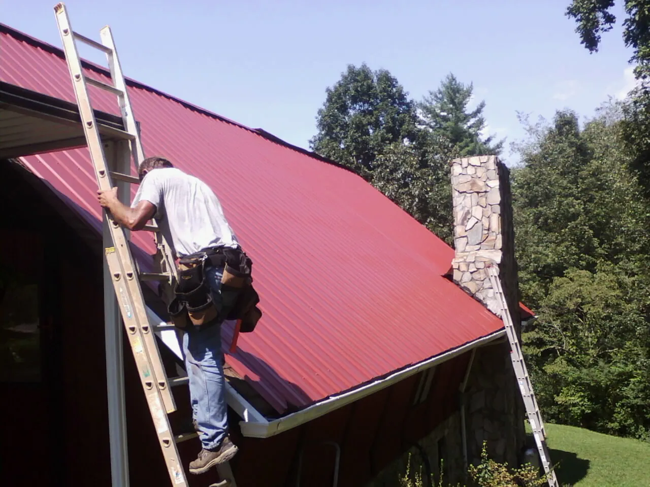 Roofer on a ladder beside a red metal roof and stone chimney — Soffit & Fascia Repair by Roofing Brothers in Mc Clellanville